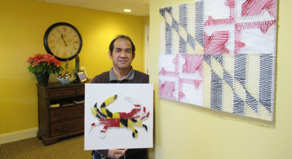 Angeles Quiozon standing next to his Maryland Flag picture while holding his Maryland flag styled crab picture; bot made from pins and thread.