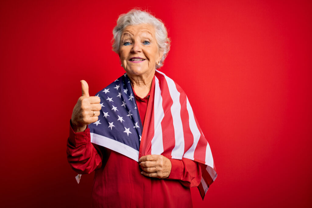 Senior veteran woman giving a thumbs up to the VA's Aid and Attendance program for senior living, Elderly woman wrapped in American flag with a thumbs up, happy senior woman wearing flag, smiling veteran with flag around shoulders