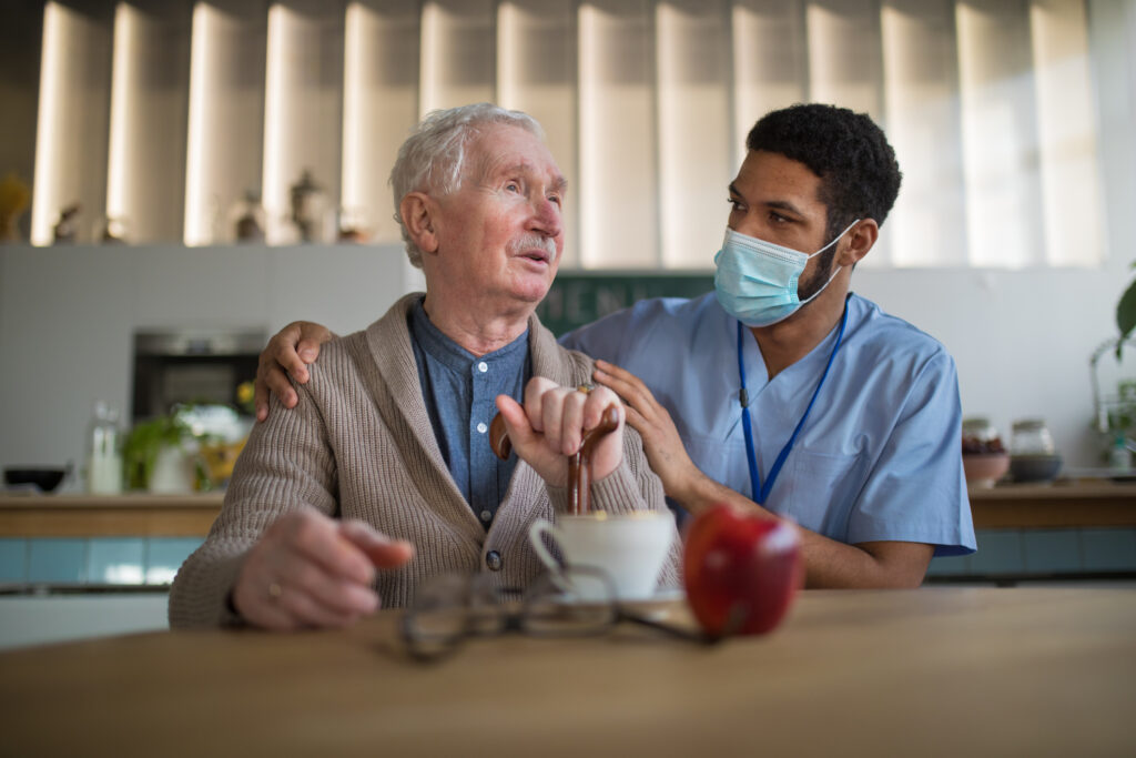 A male health care worker caring for an older man who is seated at a table.