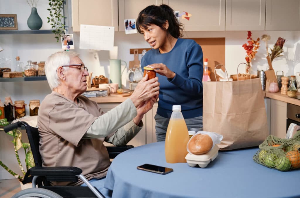 A senior man in a wheelchair receiving at-home care help with opening a jar from a caregiver.