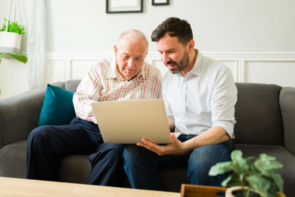 A father and son having a senior living conversation sitting on a couch while doing research on a laptop computer.