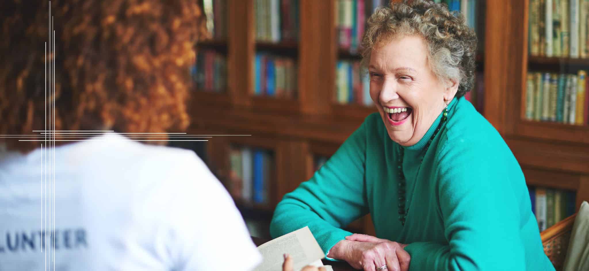 An older woman joyfully chats with a volunteer holding a book in a library setting, illustrating the meaningful connections created through volunteer opportunities.